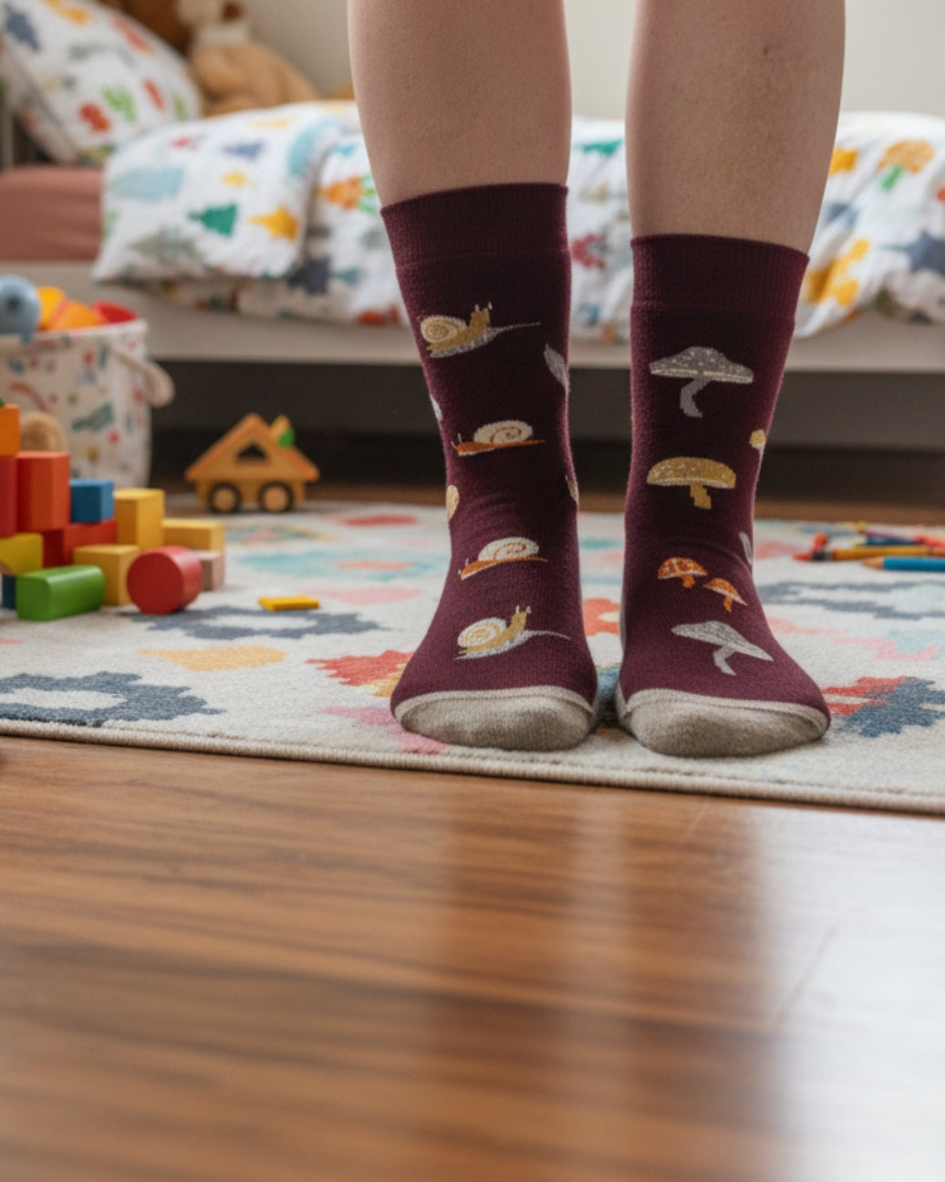 A person standing on a patterned rug in a child's bedroom, wearing burgundy crew socks with embroidered mushrooms, snails, and grasshoppers.