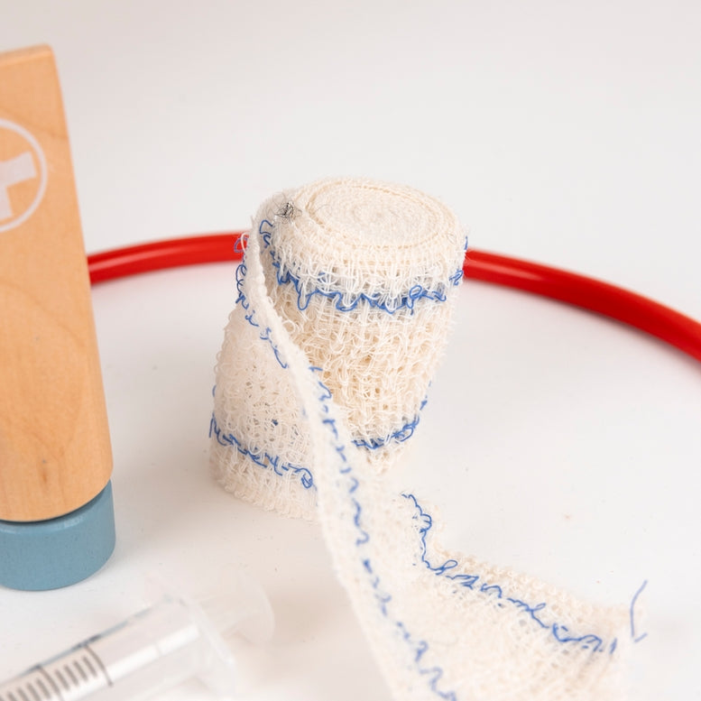 A roll of beige gauze bandage with blue stitched edges, shown unrolled on a white surface. A wooden-handled medical tool and part of a clear syringe are visible at the edges, along with a red circular prop in the background.