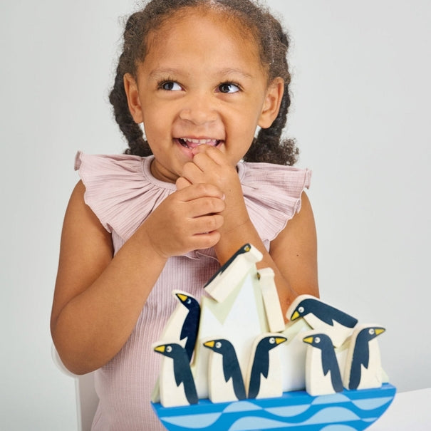 A young child with braided hair and a ruffled pink top smiling playfully while sitting next to the wooden balancing penguins and rocking iceberg toy.