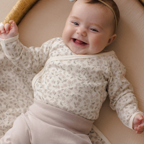 A smiling baby lying on their back in a woven bassinet, wearing a cream and beige floral long-sleeve wrap top with matching beige pants and a gray bow headband.