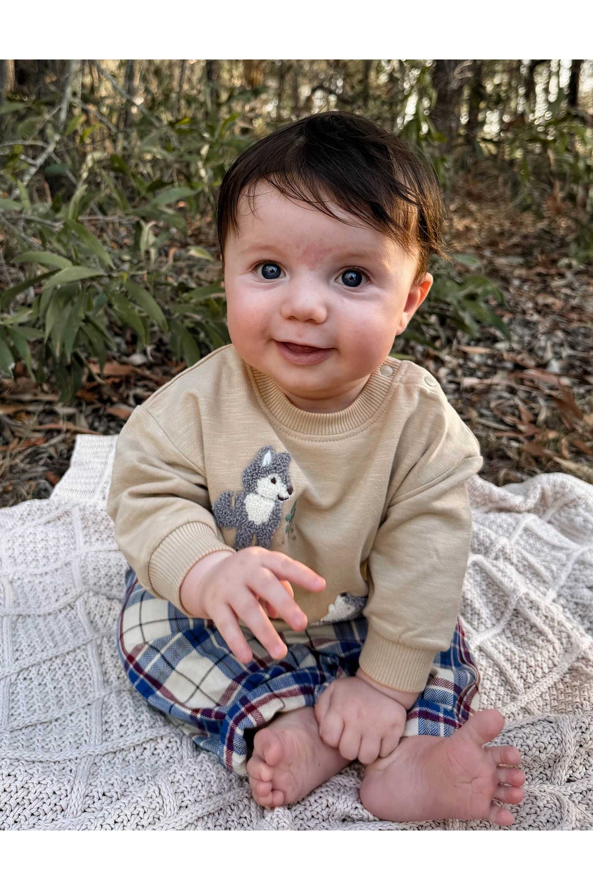 A baby sitting outdoors on a white knit blanket, wearing a beige long-sleeve shirt with a gray and white dog graphic and plaid pants.
