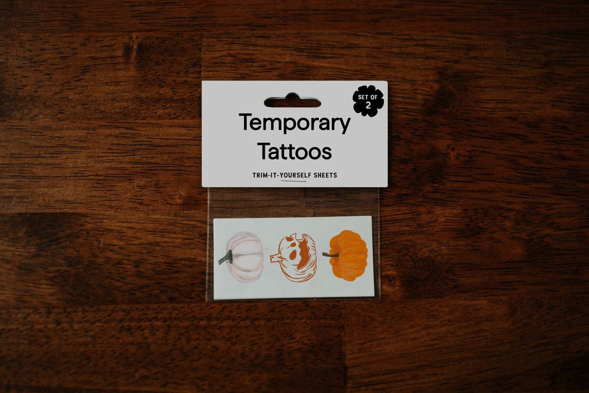 Pack of temporary tattoos on a dark wood background. The white card label reads 'Temporary Tattoos' and shows three pumpkin designs on the sheet below.