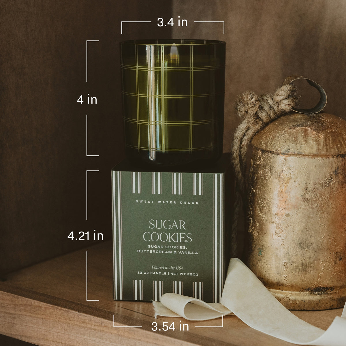 A candle in a dark green glass jar with a plaid pattern sits on top of its matching box. White measurement lines and text indicate the candle is 4 inches tall, the box is 4.