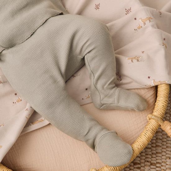 A close-up of a baby's legs wearing muted sage green waffle-knit booties, resting on a patterned cream muslin blanket inside a woven rattan bassinet.