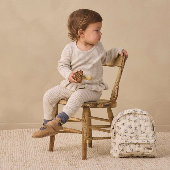A toddler sits on a rustic wooden chair in a studio setting, wearing a cream ribbed long-sleeve top and matching cream pants. A small floral backpack rests beside the chair against a warm beige backdrop.