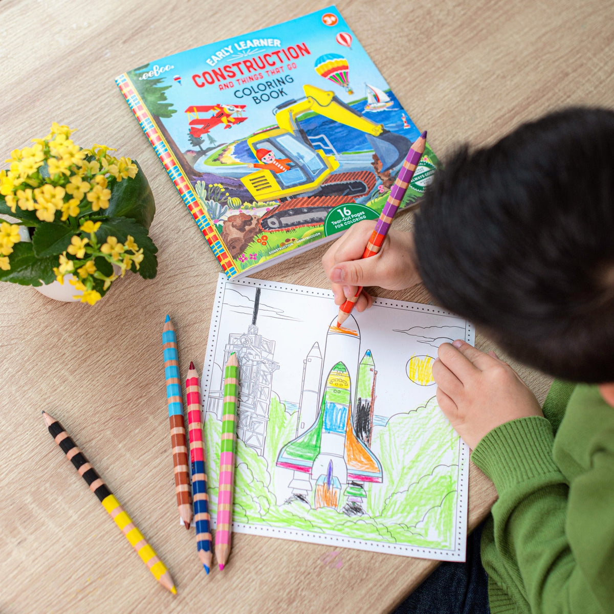 Child coloring a rocket ship with a construction-themed coloring book and crayons on a wooden table.