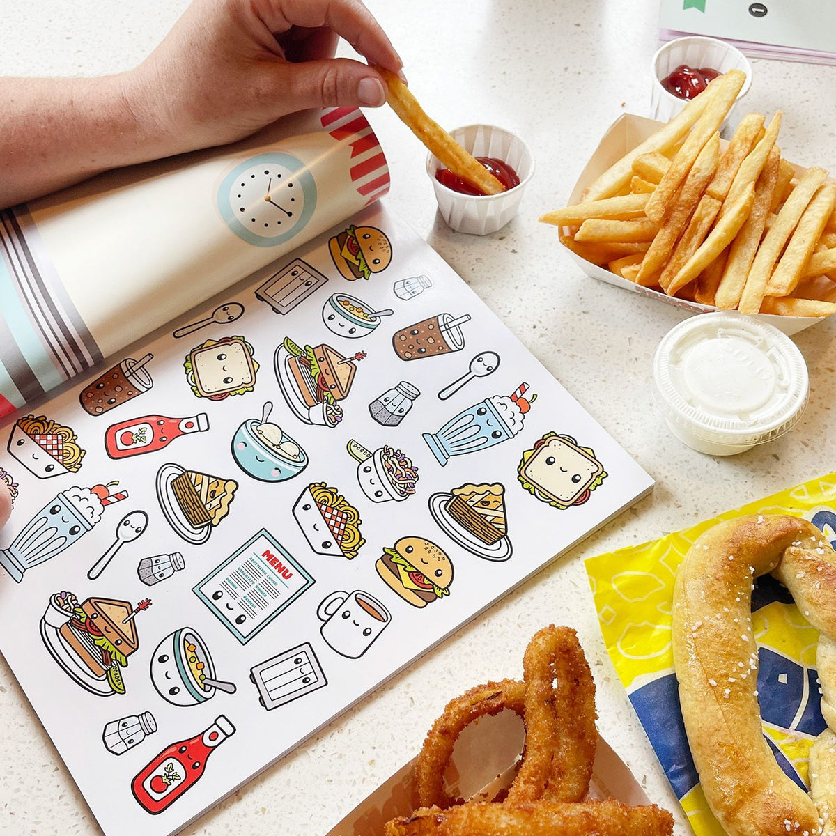 Coloring book with food illustrations next to snacks on a table.