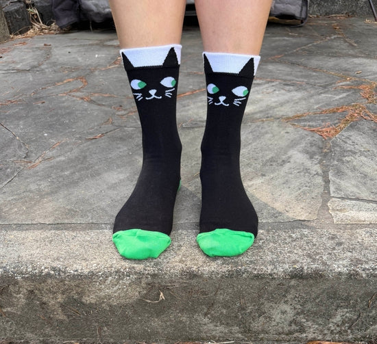 Black crew socks with a cartoon cat face design and bright green toes, worn on feet standing on a stone surface.