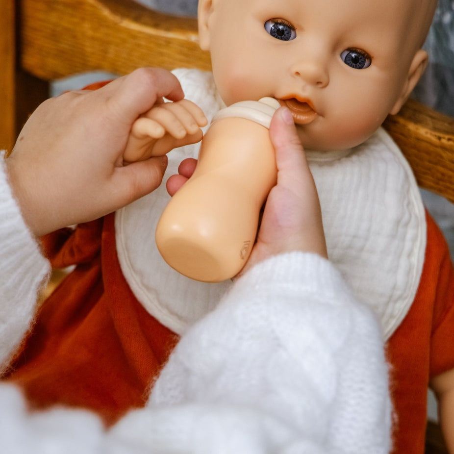 A close-up of a baby feeding doll bottle scene, showing a person's hands holding a beige baby bottle to a doll's mouth. The doll has a bald head, realistic skin-toned face, blue eyes, a white bib, and an orange-red outfit, seated in a wooden chair.
