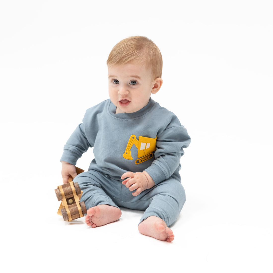 A baby sitting on a white studio background wearing a light blue one-piece sweatshirt and pants set with a yellow excavator graphic on the chest, holding a small wooden toy.