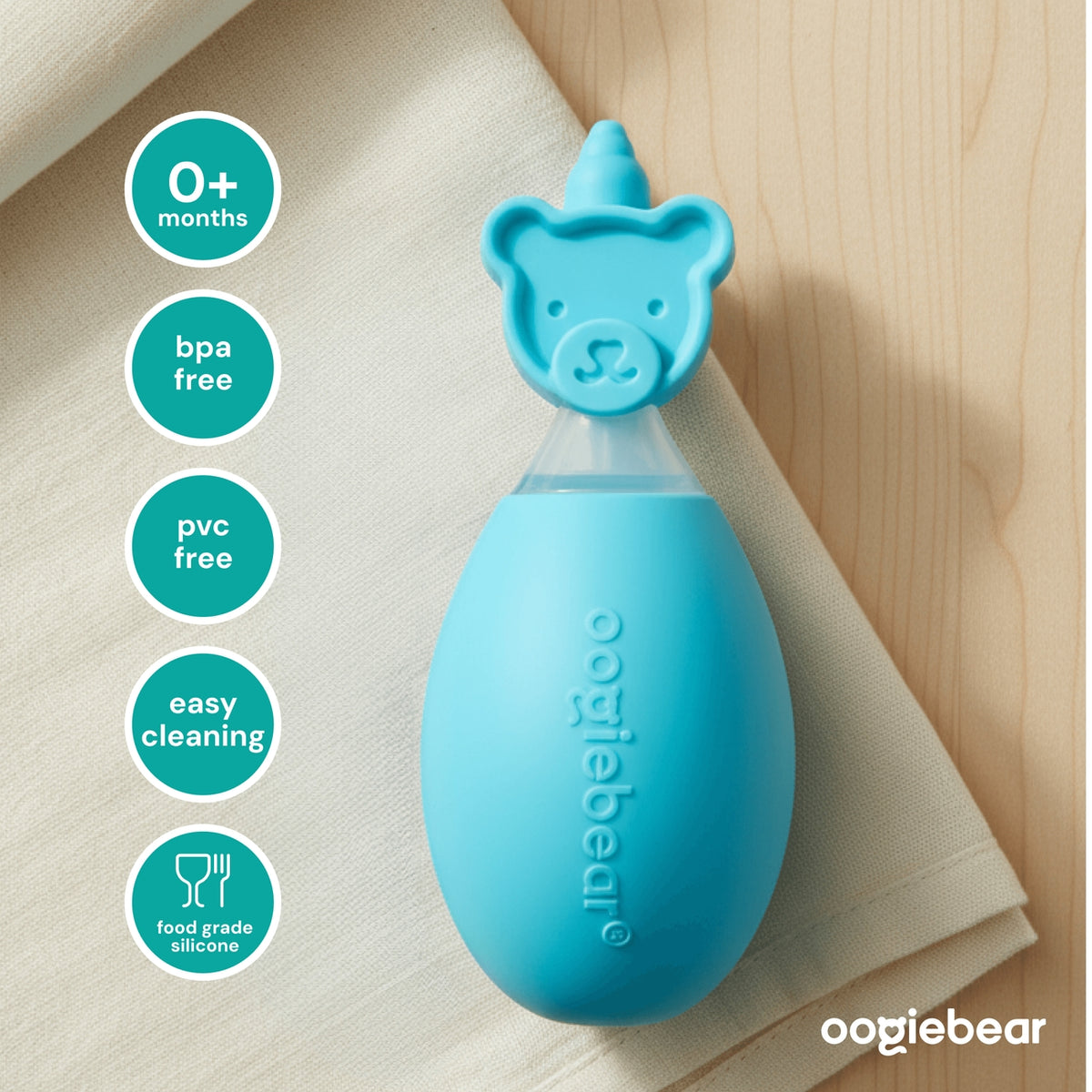 A light blue Oogiebear baby bottle with a bear-shaped cap, styled on a beige cloth over a light wood background. On the left are five teal circular badges highlighting product features: 0+ months, bpa free, pvc free, easy cleaning, and food grade silicone.