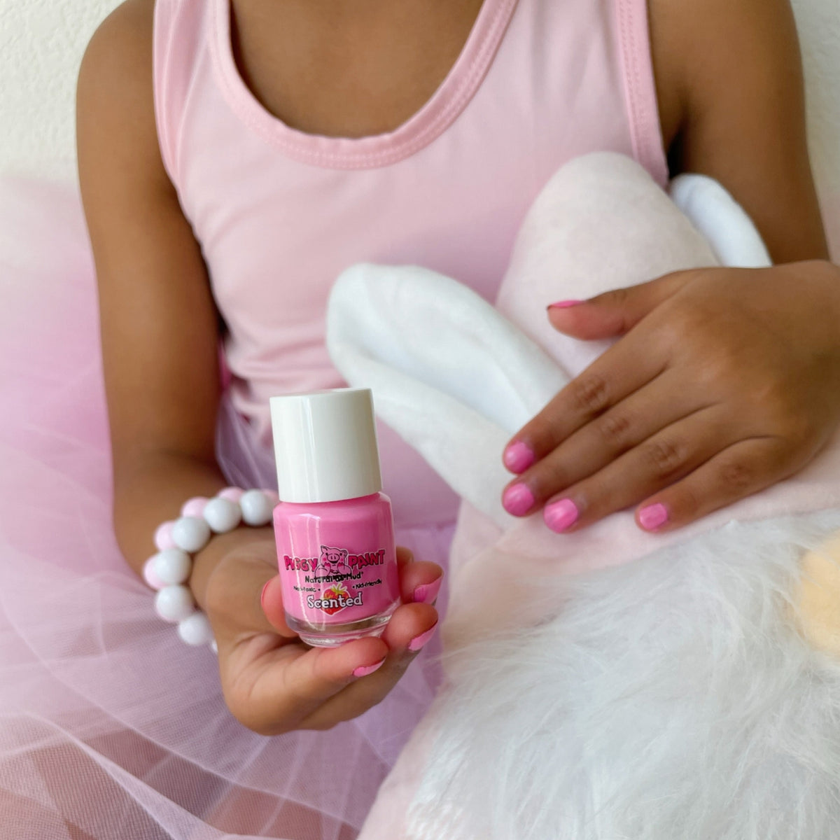 Child in pink dress holding a bottle of pink nail polish with a white cap.