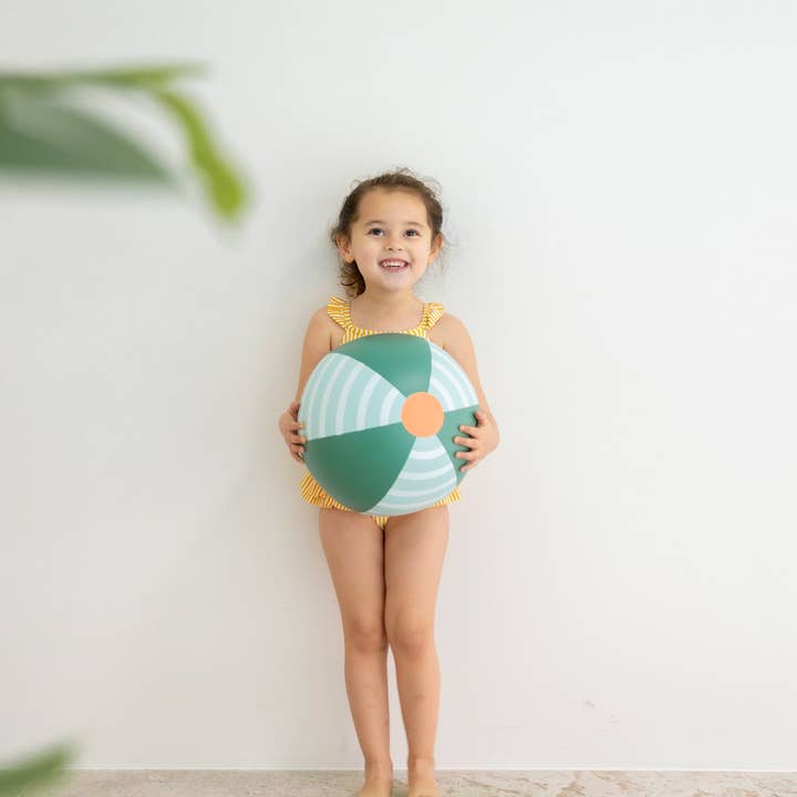 A young child smiling while wearing a yellow striped one-piece swimsuit, standing indoors next to a green and light blue striped beach ball.