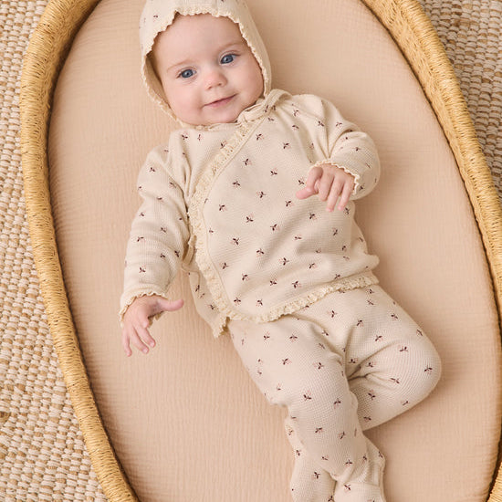 A baby wearing a cream-colored footed sleeper with a matching bonnet, decorated with tiny brown cross patterns, lying on a beige cushion inside a woven oval basket.