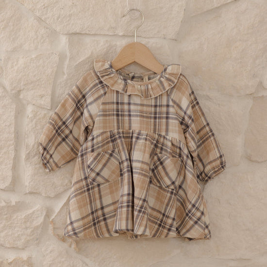 A toddler girl's long-sleeve plaid dress in beige, brown, and black, hanging on a wooden hanger against a rustic stone wall.