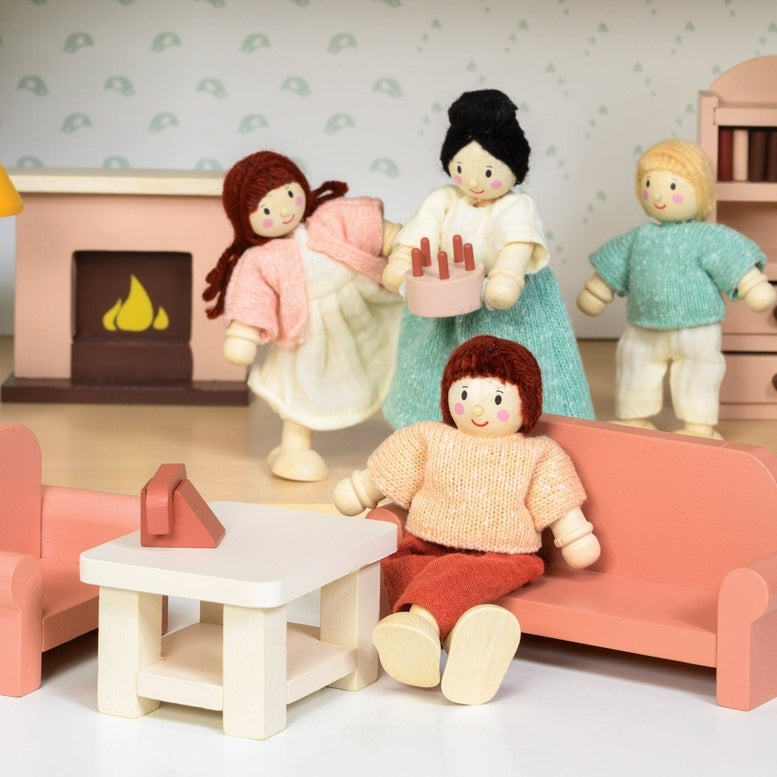 A miniature wooden dollhouse room interior featuring a wooden toy figure standing next to a coral-pink sofa and armchair set, a coffee table with a tiny red telephone, a pale pink decorative fireplace, a yellow floor lamp, and a natural wood bookcase against a patterned wallpaper background.
