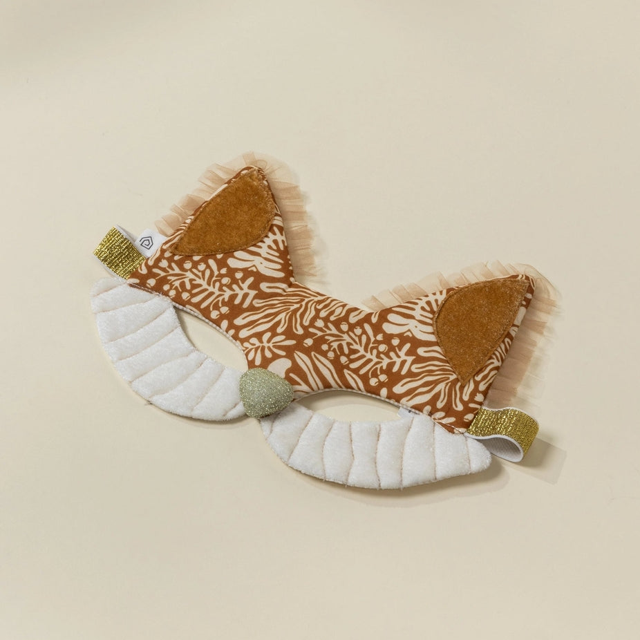 A plush fox-themed baby headband with patterned brown and white fabric ears, white padded cheeks, and small gold glitter accents, displayed on a light beige background.