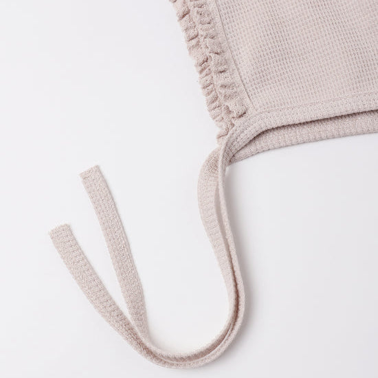 Close-up of a light beige knit garment with a ruffled trim and matching tie straps on a plain white background.