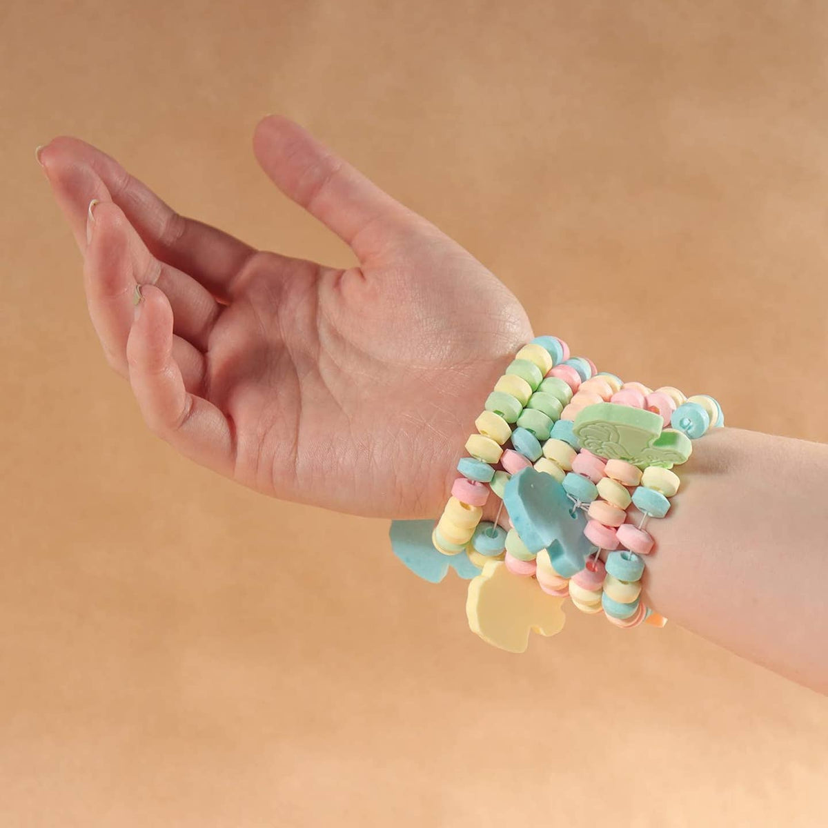A close-up of a person's hand and wrist wearing a stack of pastel candy-themed bracelets, including pastel beads and elephant-shaped charms, against a solid tan background.