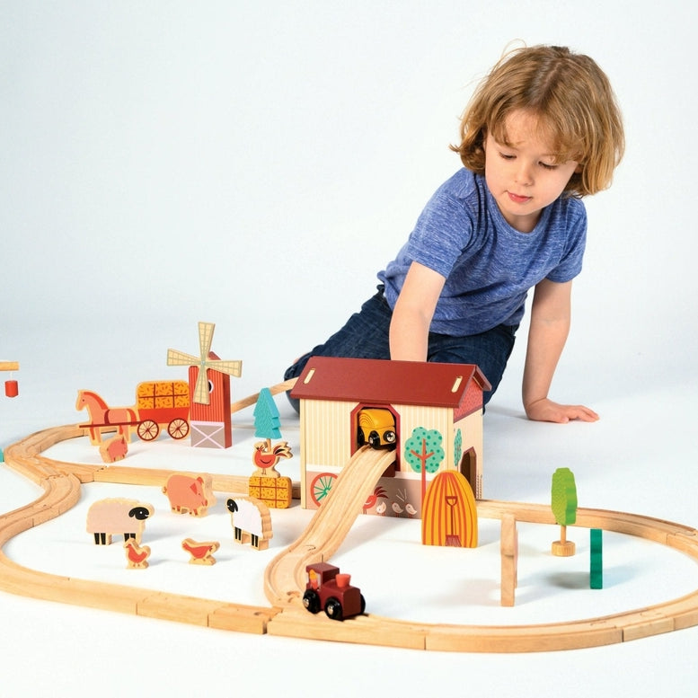 A young child plays on the floor with a wooden farmyard-themed train set, arranging the tracks, red barn, and windmill while placing a small train in motion.