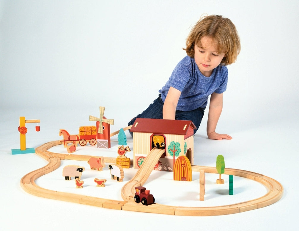 A young child plays on the floor with a wooden farmyard-themed train set, arranging the tracks, red barn, and windmill while placing a small train in motion.