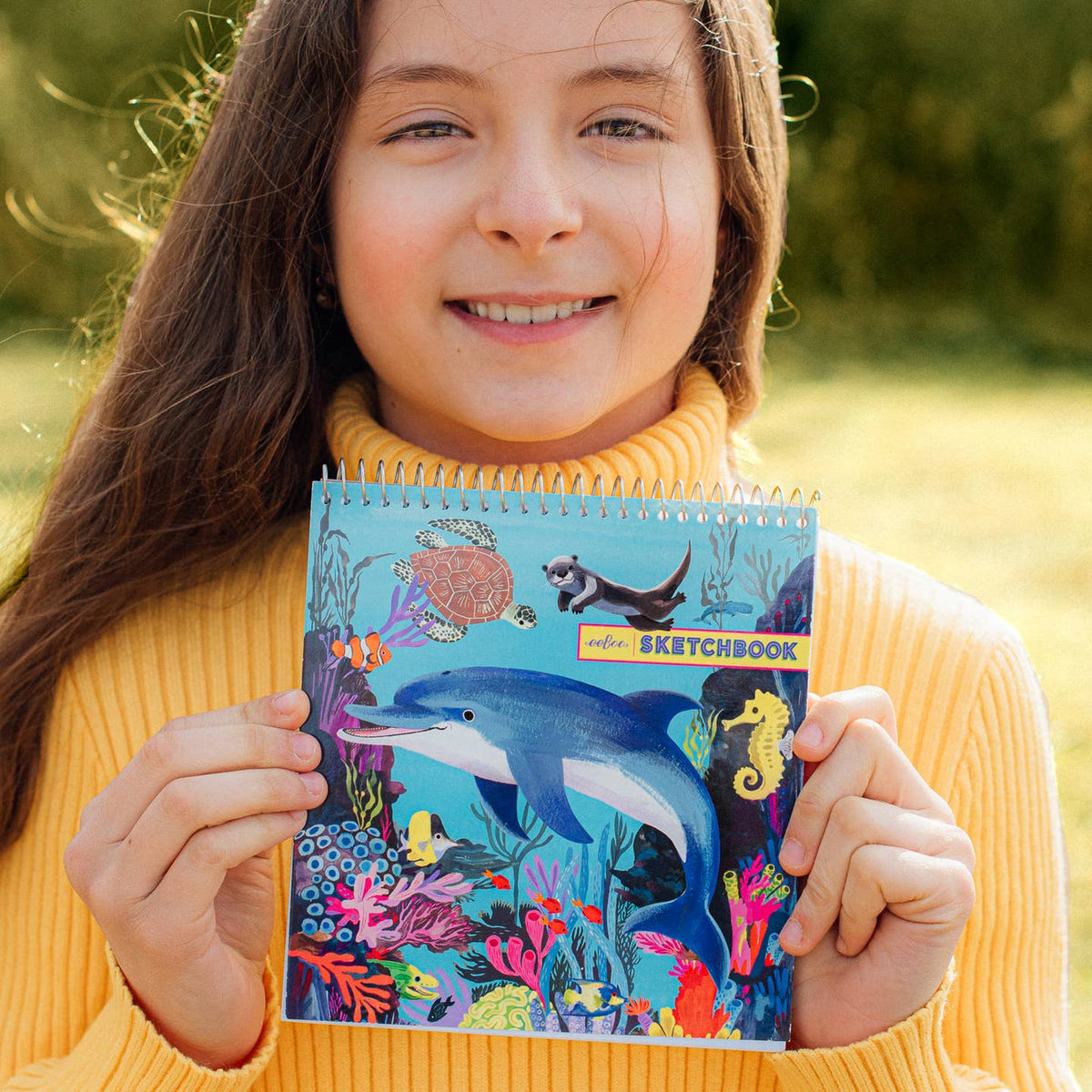 Girl holding a colorful sketchbook with an underwater design, outdoors.