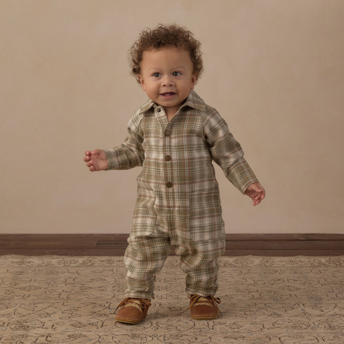 A toddler standing and wearing a long-sleeve plaid romper in muted green, beige, and brown tones, paired with brown shoes, against a warm beige studio backdrop.