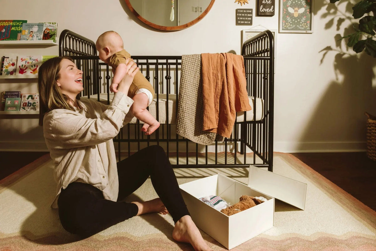 Woman holding a baby in a nursery with a crib and toys on the floor.