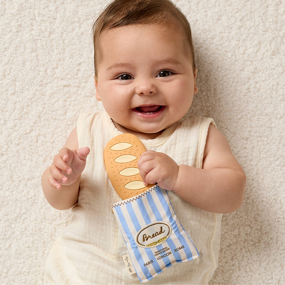 Baby holding a toy loaf of bread and package on a soft surface