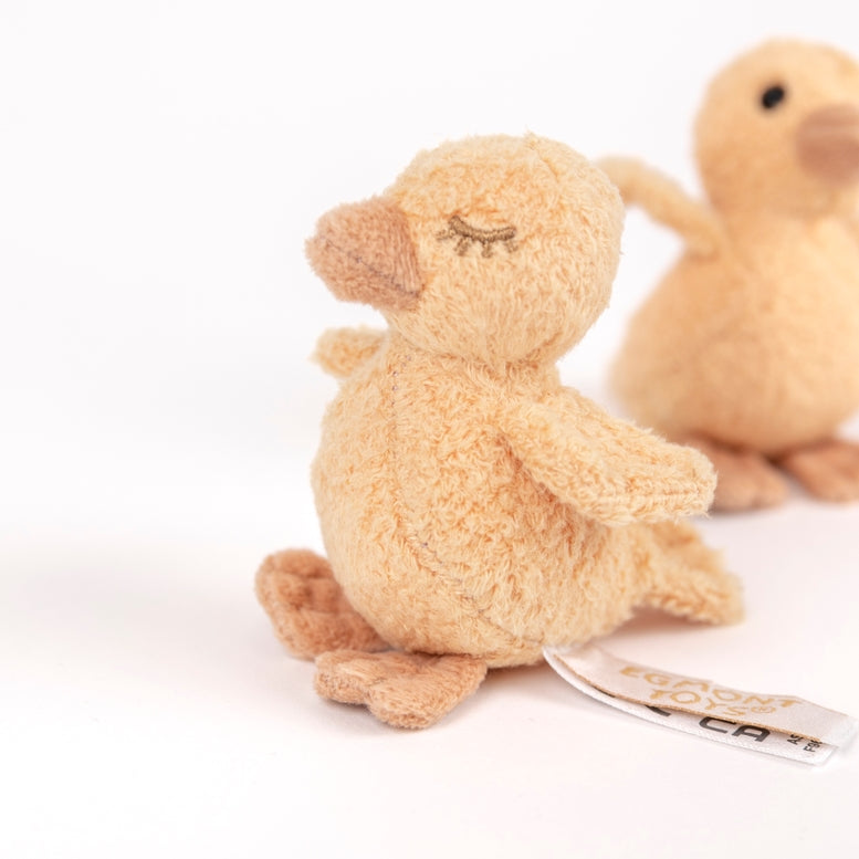 A small plush stuffed duck in a warm beige/tan color, shown from the back with its embroidered closed eyes and a small paper tag attached. Another similar plush duck is softly blurred in the background on a clean white studio surface.