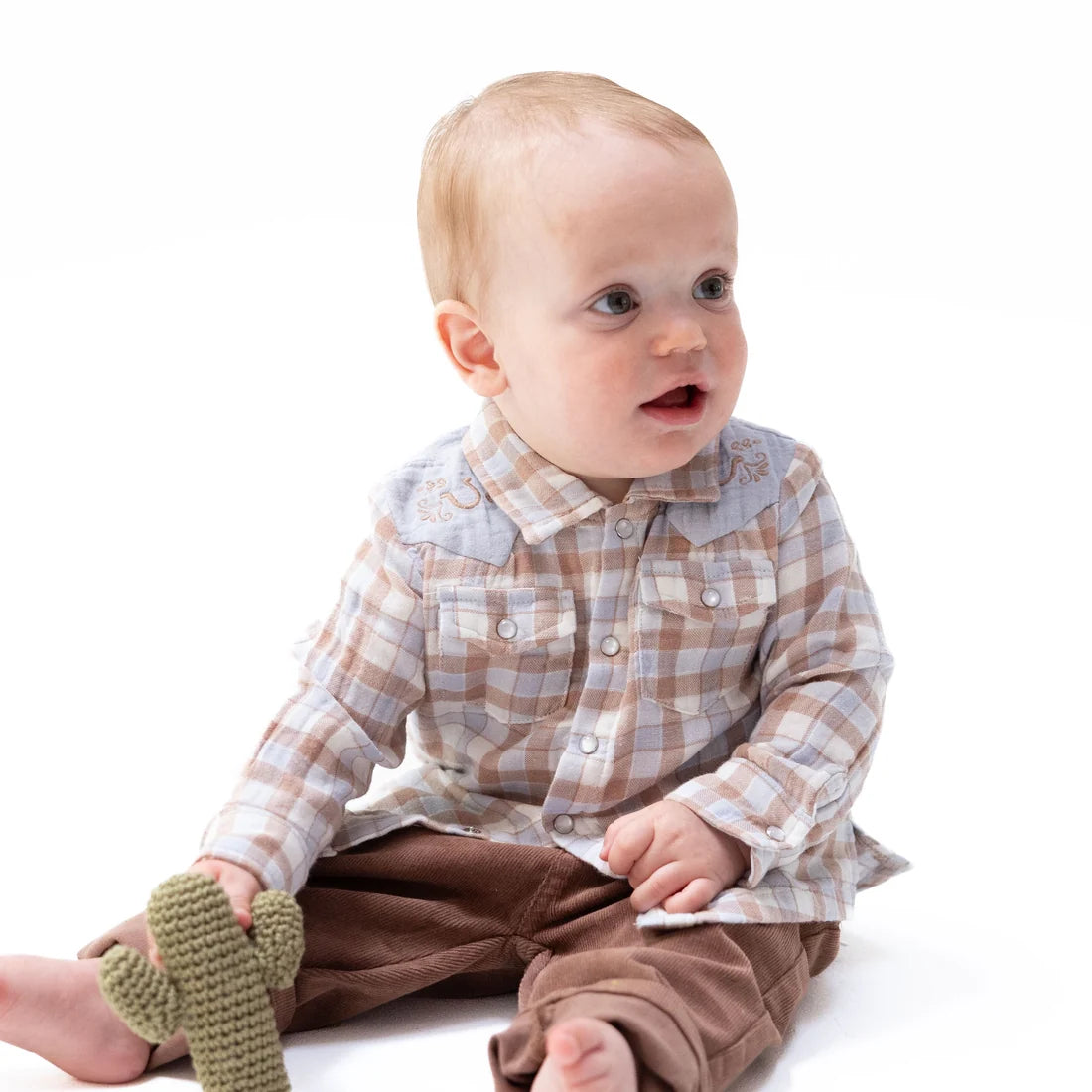 A baby sitting on a white background wearing a brown and beige plaid long-sleeve shirt, brown pants, and holding a green cactus-shaped knit toy.