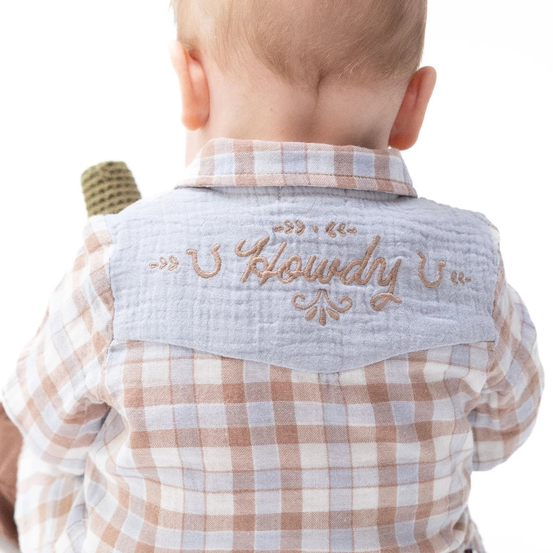 Back view of a baby wearing a light blue muslin neck gaiter with 'Howdy' embroidered in tan script, layered over a beige and white checkered shirt.