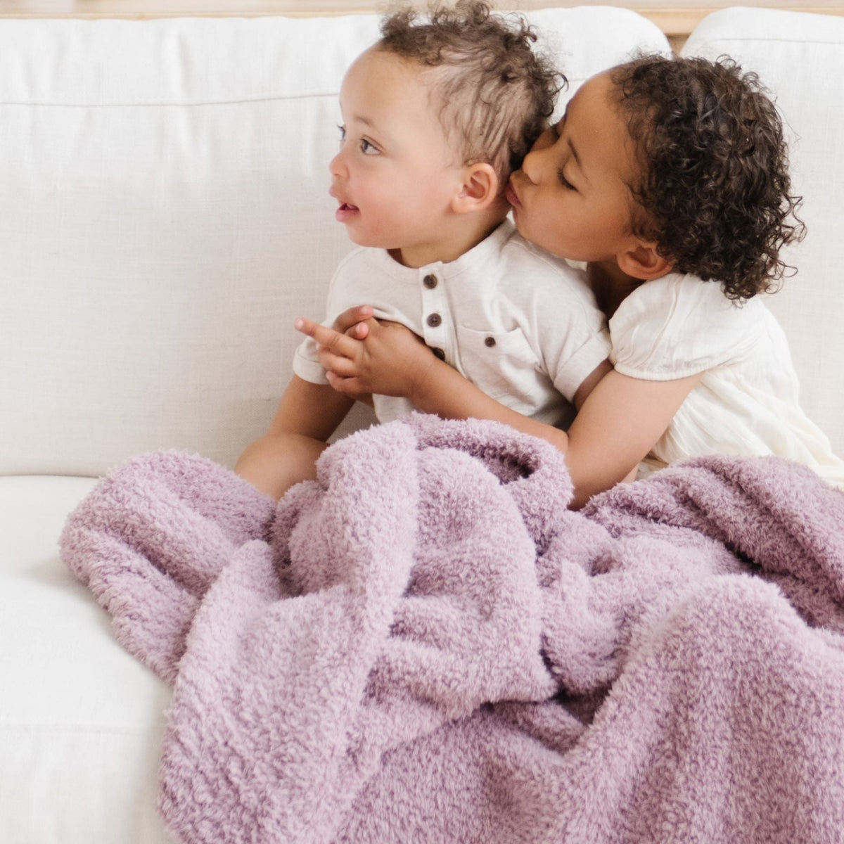 Two children sitting on a couch under a purple blanket