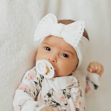 A baby lying on a soft white blanket, wearing a white lace-trimmed bow headband and a white floral long-sleeve romper.