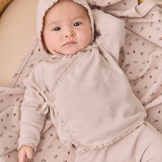 A baby wearing a light beige waffle-knit bonnet and matching long-sleeve romper, lying on a floral muslin blanket in a woven basket.