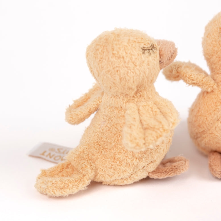 A close-up of a soft, textured plush bunny in a warm beige/tan color, shown from the back with its small fabric tag visible. Part of another similar plush toy is cropped on the right side against a clean white background.