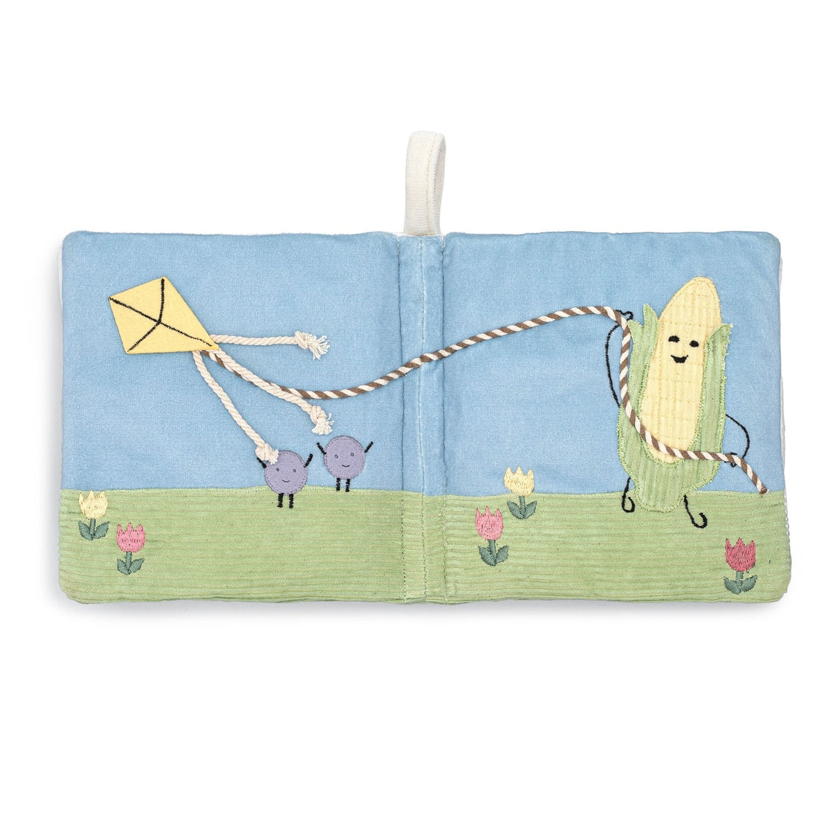 A square fabric book or pouch with a stitched outdoor scene on the cover, featuring a blue sky, green grass, flowers, a yellow kite, and a smiling corn character holding a rope. A white hanging loop is attached at the top center.