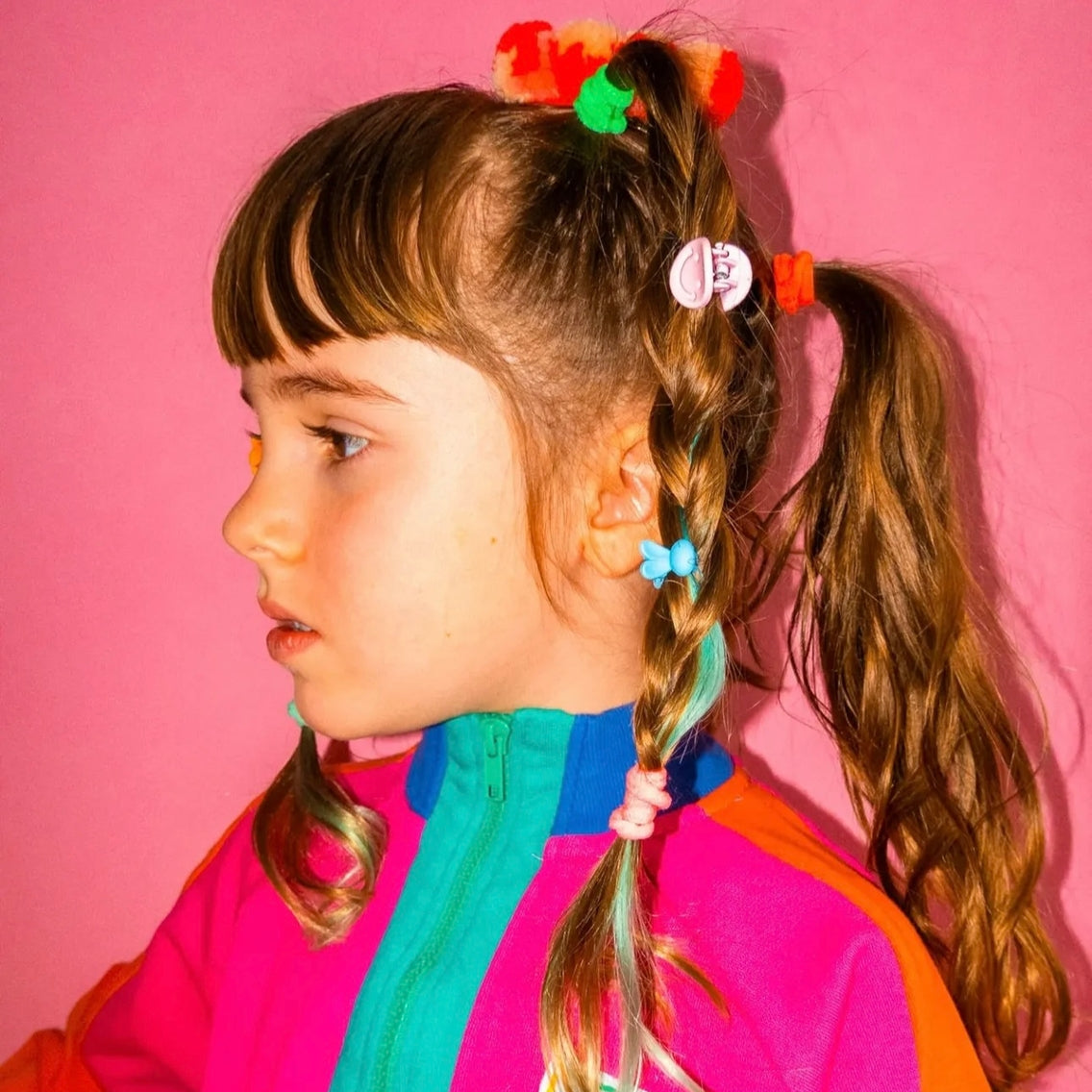 Child with colorful hair accessories and a multicolored jacket against a pink background