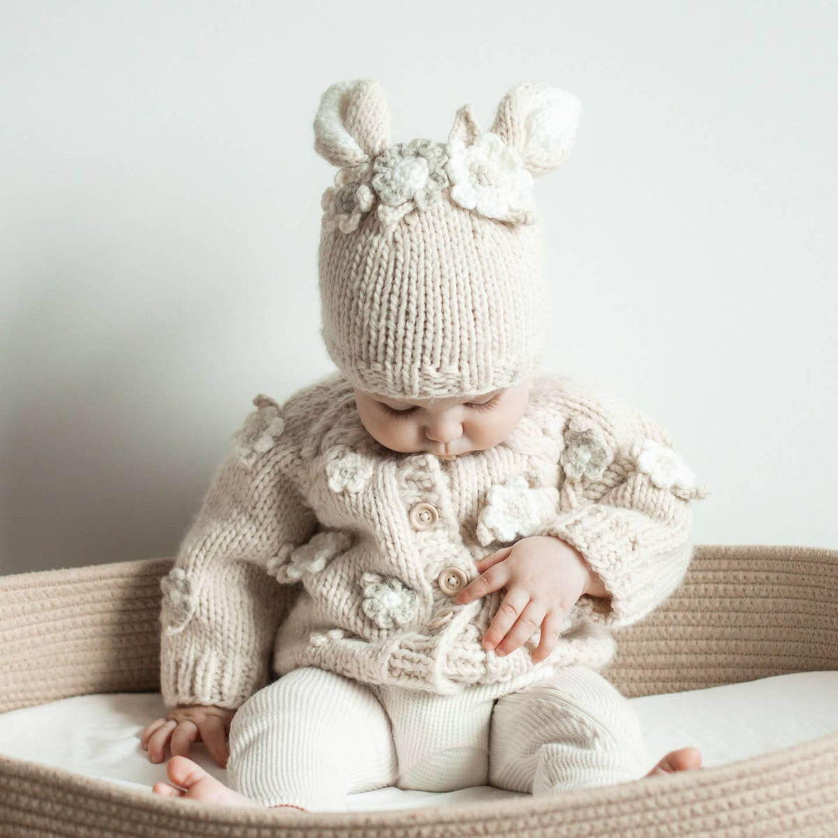 Baby in a knitted outfit with floral details sitting in a crib.