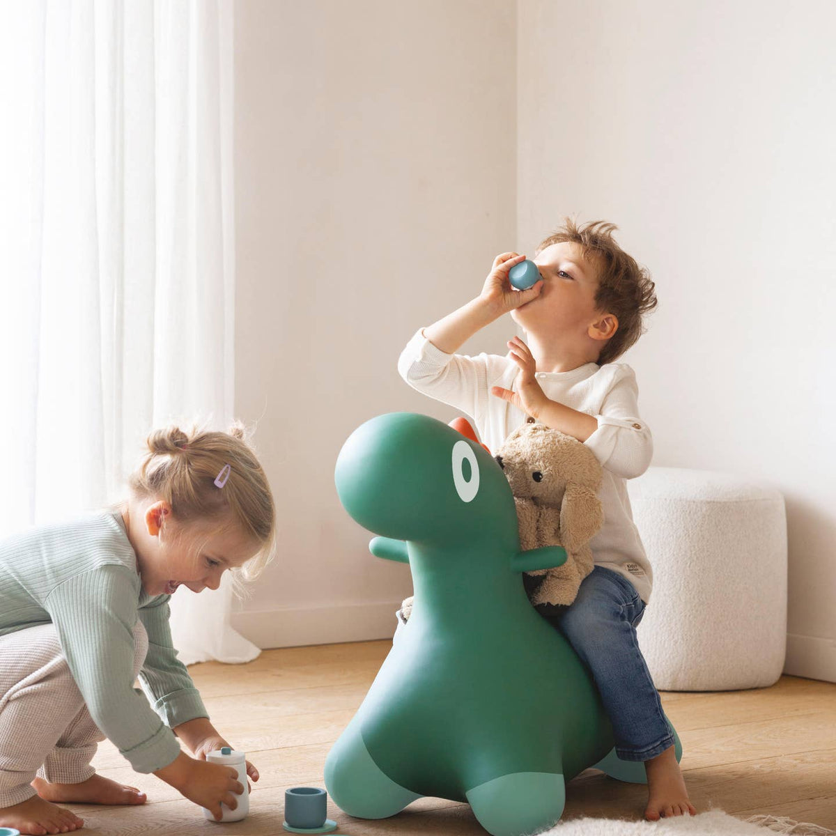 Two children playing with a green dinosaur-shaped toy in a room with light-colored walls and wooden floor.