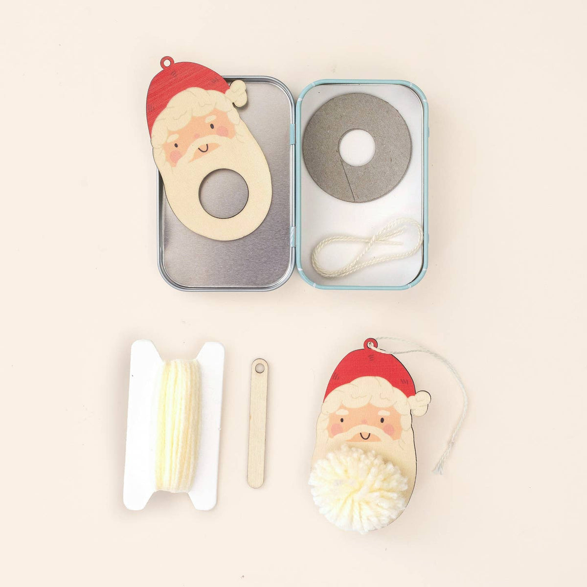 A Christmas-themed wooden ornament craft kit shown in its open metal tin with included pieces laid out below on a light beige background. The kit contains a Santa face ornament, a gray wooden ring, a white pom-pom, a spool of white thread, and small wooden tools.