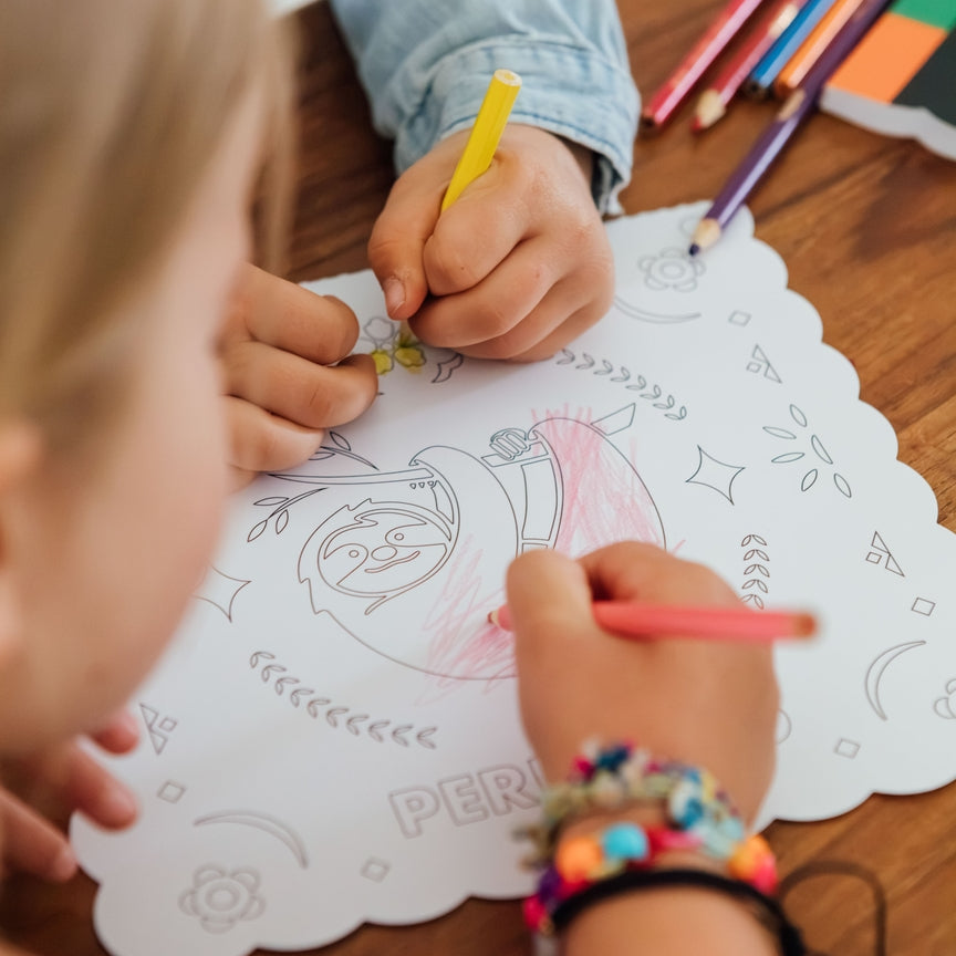 A close-up lifestyle shot of children coloring on a white scalloped paper sheet with a cartoon character. One child is drawing with a yellow pencil, and another is drawing with a pink pencil.