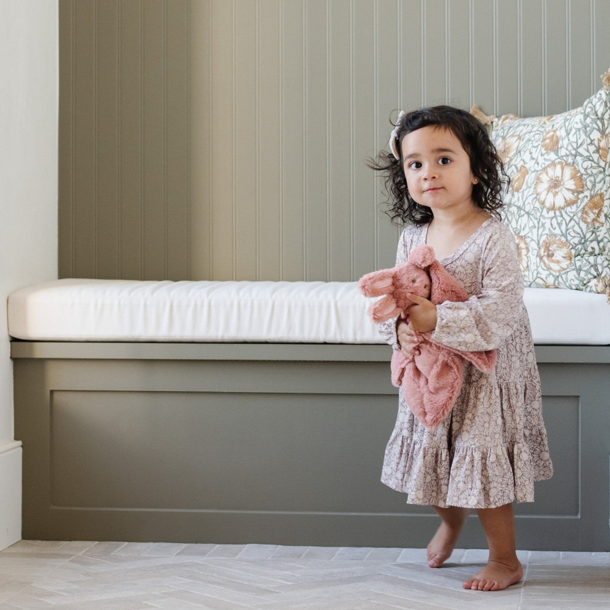 A toddler girl with curly dark hair stands barefoot in a nursery, wearing a long-sleeve floral baby dress and holding a pink plush bunny toy. She is positioned in front of a built-in bench with a white cushion and a patterned pillow against a beige paneled wall.
