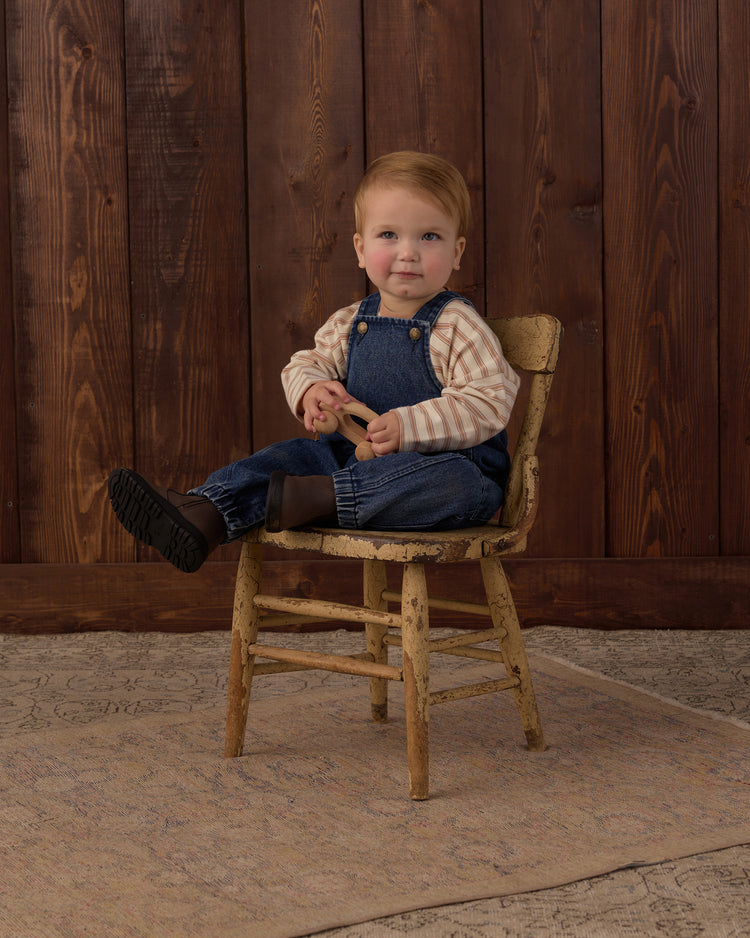 A toddler with light hair sits on a rustic wooden chair against a dark wood plank wall. The child is wearing blue denim overalls over a beige and white striped long-sleeve shirt, paired with dark socks and shoes, holding a small wooden toy.