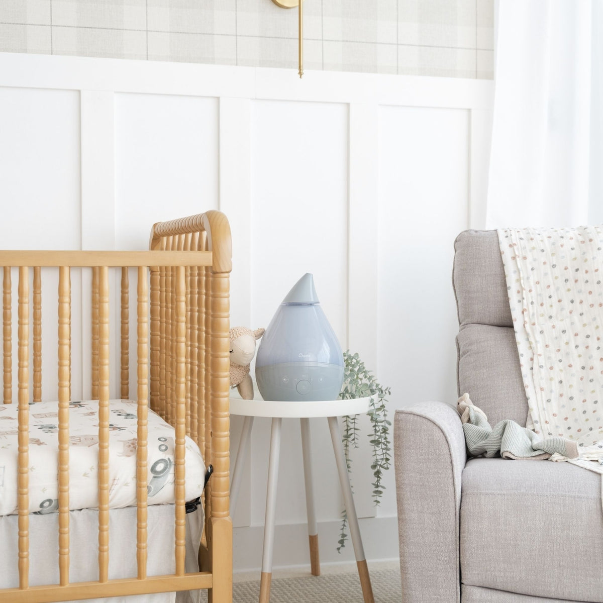 A styled nursery corner featuring a light wood spindle crib on the left, a small white side table with a matte blue diffuser in the center, and a light gray fabric armchair on the right against a white paneled wall.