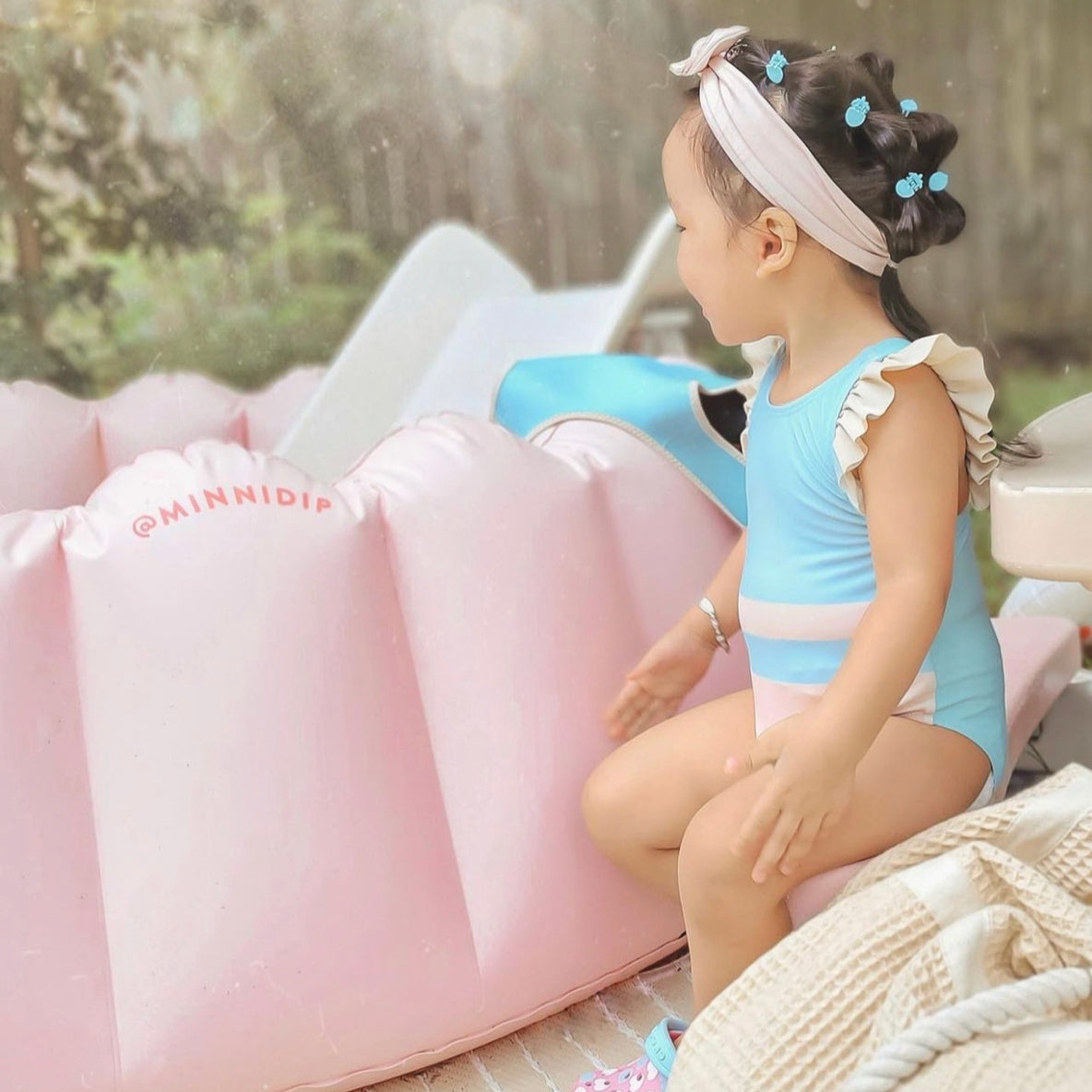 Child in a blue swimsuit sitting on a pink inflatable chair outdoors.