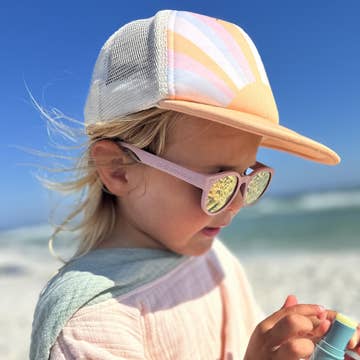A young child wearing a pastel striped trucker hat and pink sunglasses with mirrored lenses, standing outdoors near the beach under a blue sky.