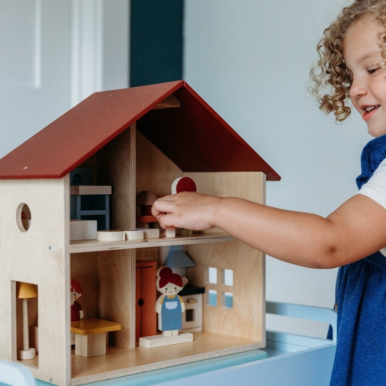 A young child with curly brown hair interacting with an open-faced wooden dollhouse by Mentari, placing miniature furniture and small figurines inside the rooms.