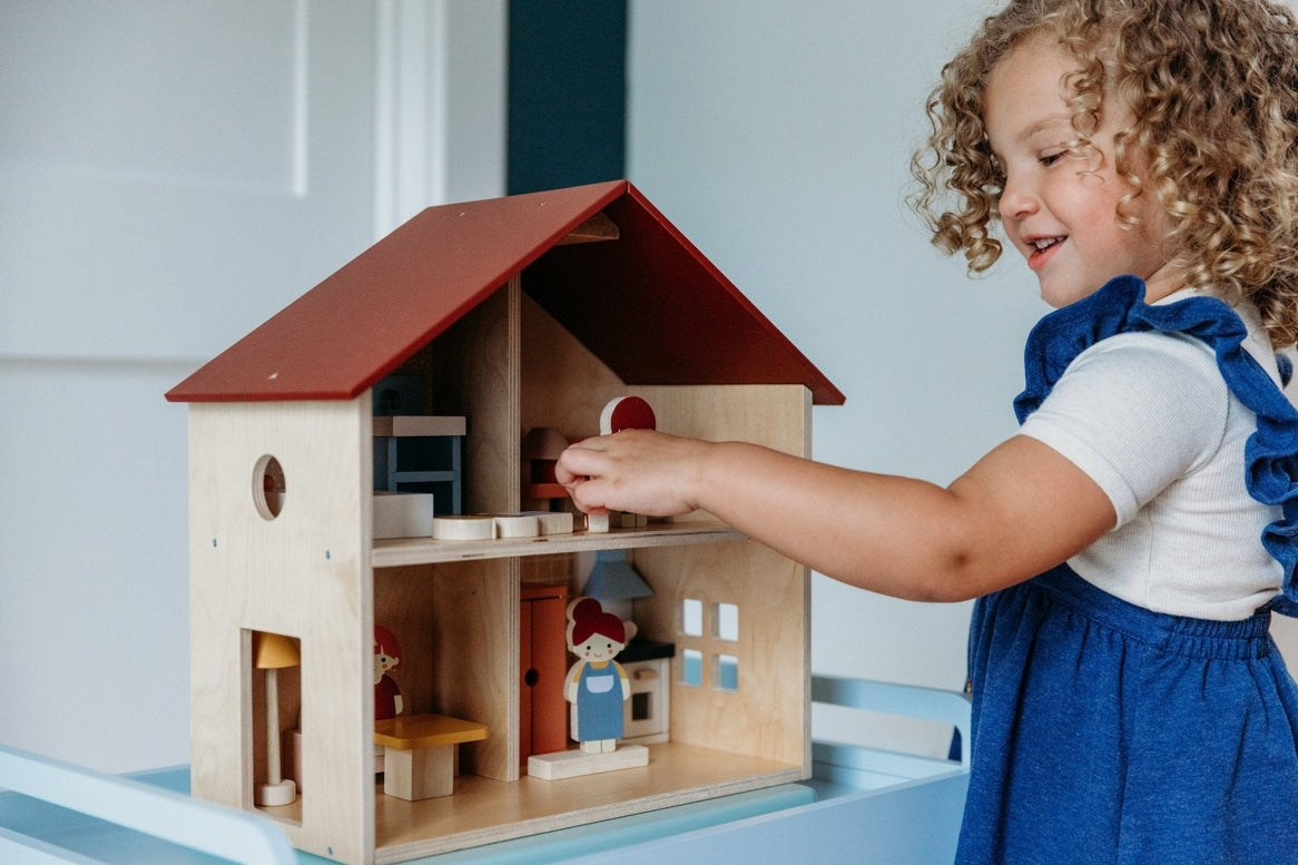 A young child with curly brown hair interacting with an open-faced wooden dollhouse by Mentari, placing miniature furniture and small figurines inside the rooms.