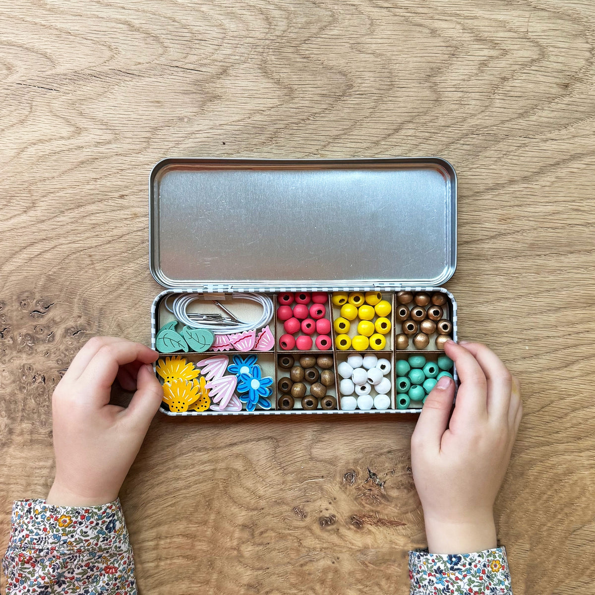 Child's hands interacting with a small metal box filled with colorful beads and craft supplies on a wooden surface.